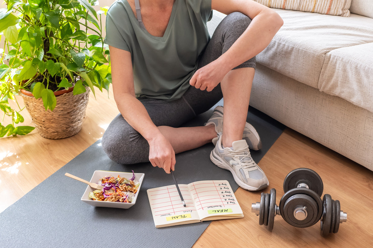 Woman sitting on an exercise mat at home while planning healthy habits in a notebook.